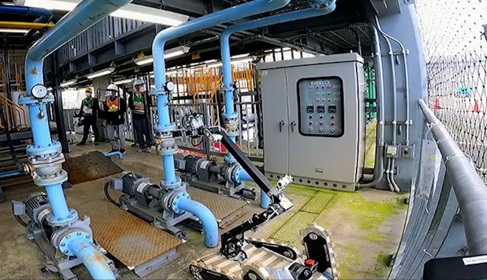 産業プラントの配管とポンプ。手前にクラウラ型点検ロボット、奥にヘルメットを着用した三名の作業者。